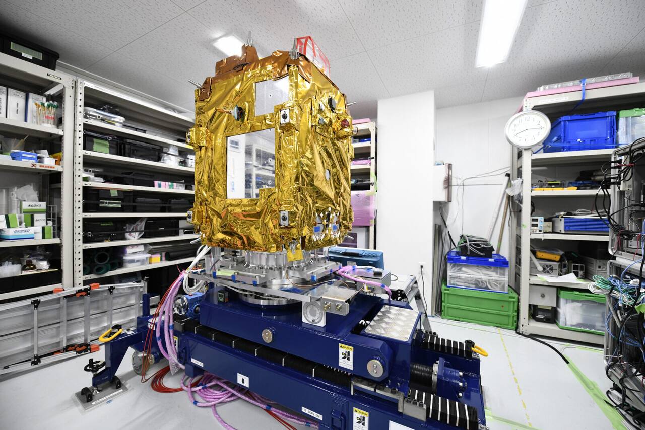 ADRAS-J on a handling and vibration fixture in the cleanroom before launch. The bus is wrapped in multi-layer insulation, the two-panel solar wings are stowed against the side panels, and the rendezvous payload apertures are the cutouts on the near face.