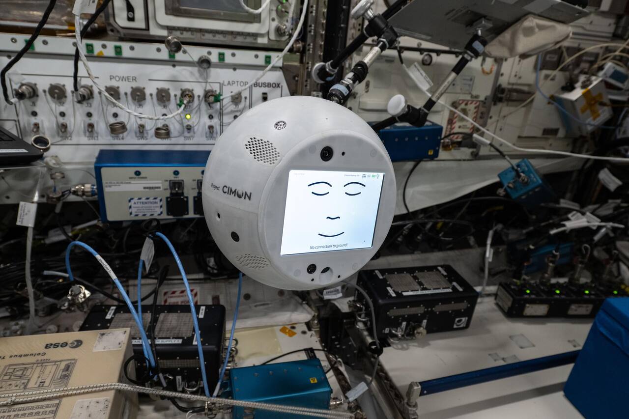 CIMON-2 free-flying in the Columbus module on 18 July 2025, just after activation. The 32 cm sphere has no external moving parts; the twelve fans that provide both translation and attitude control are internal, and the small circular apertures above and below the display are camera and microphone ports. The LCD carries the animated face used as the status display, here reporting No connection to ground, which is the failure mode implied by running the language understanding on Earth rather than onboard