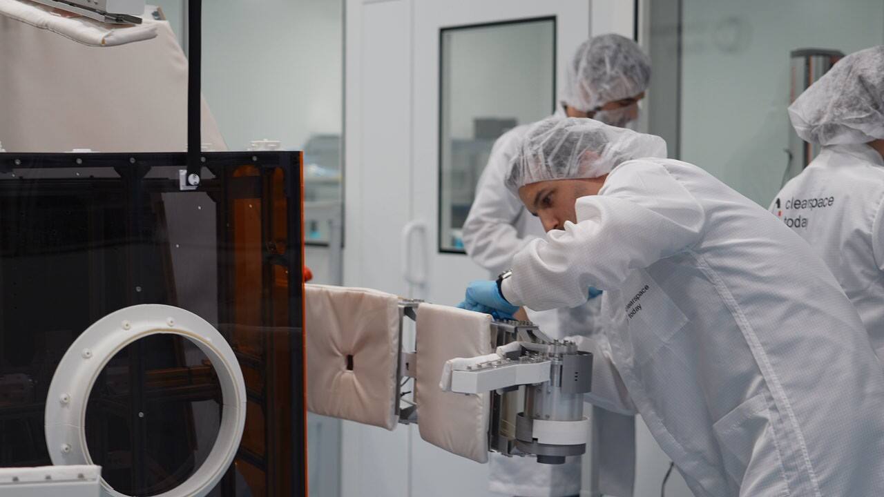 A ClearSpace capture arm being mounted to a spacecraft panel in the ClearSpace cleanroom. The arm segments are covered in thermal blanketing; the joint actuator and its gear housing are exposed at the elbow, and the circular fitting on the panel at left is the arm root interface.