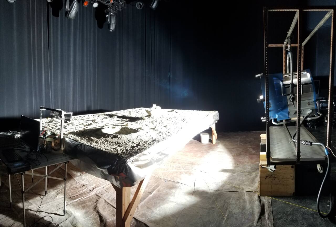 Perception testbed at the Carnegie Mellon Robotics Institute under an ARRI Arrimax 18/12 HMI head mounted at bed height on a rolling stand. The lamp is set near grazing incidence to reproduce the near-tangential illumination of the lunar pole; blackout curtains on the walls and floor suppress diffuse return so that shadows stay close to black.