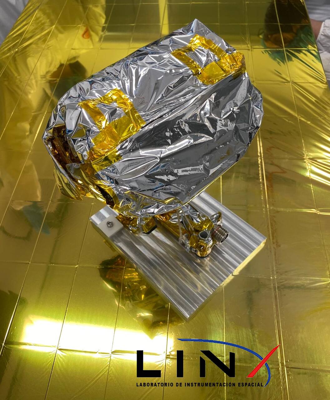 The Colmena-1 flight payload wrapped in multi-layer insulation on its aluminum lander interface plate. The closed bucket and shroud that hold the five micro-rovers sit on the catapult arms above the plate, and the electrical connector to the Peregrine lander is at the plate's forward edge