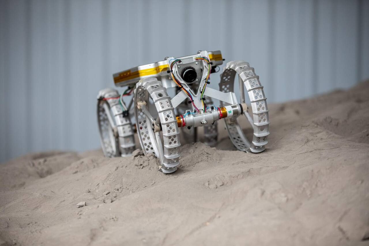 CubeRover mobility test article climbing a regolith simulant slope in the Granular Mechanics and Regolith Operations laboratory at NASA KSC, 30 June 2022. Four independently actuated wheels of perforated aluminum with integral grousers are carried on stub axles from a central boom; the forward camera housing is visible on the A-frame above the front axle, and the chassis is stripped to the mobility system for drawbar pull, slope and point-turn measurement