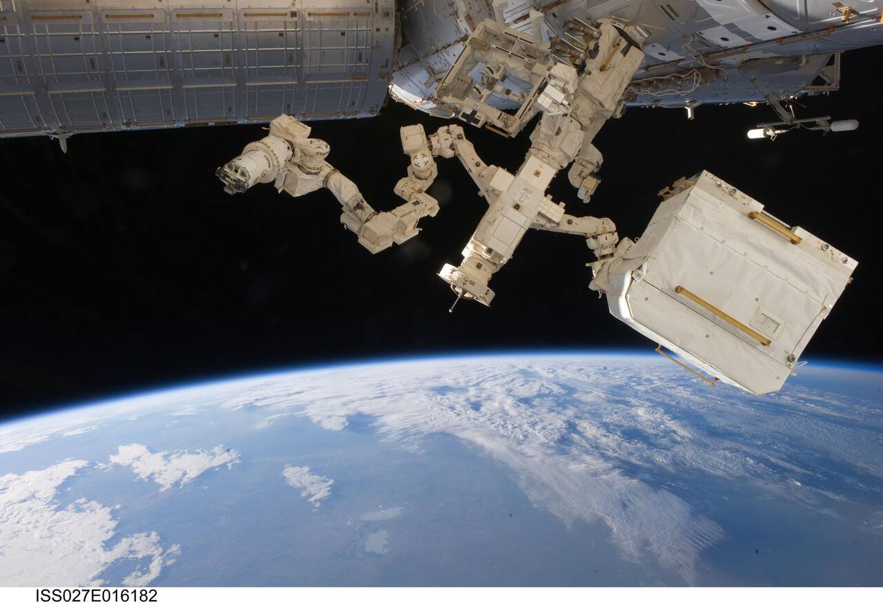Dextre holding a Cargo Transport Container on the International Space Station, 17 April 2011. Both seven-jointed arms are extended from the body, which carries the single roll joint at the waist and is anchored at top to a grapple fixture on the station; the Orbital Replacement Unit Tool Changeout Mechanism at each arm tip is the cylindrical unit from which the container is suspended at right. The container is a payload of the class Dextre exchanges in place of a spacewalk