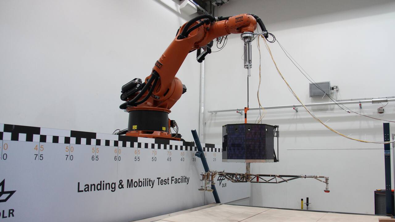 A KUKA KR500 on its linear rail holds a Philae model over the LAMA soil bed through the weight offloading suspension visible at the flange, with the lander's three-legged gear extended and a photogrammetric scale board along the bin wall.