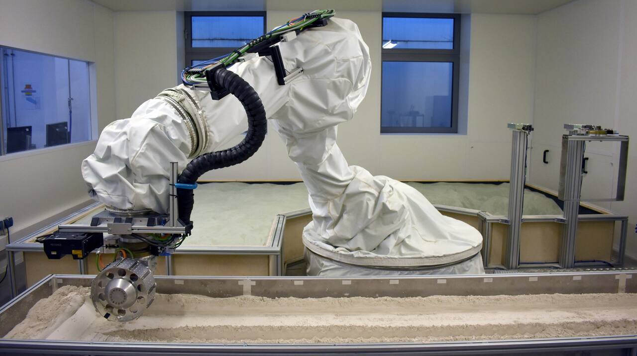 The TROLL rig: a KUKA KR210 industrial arm inside a white dust-protective jacket, holding the wheel drive unit down into the narrow front soil bin, with a Keyence laser profile scanner mounted ahead of the wheel and the larger fixed bin behind the robot.