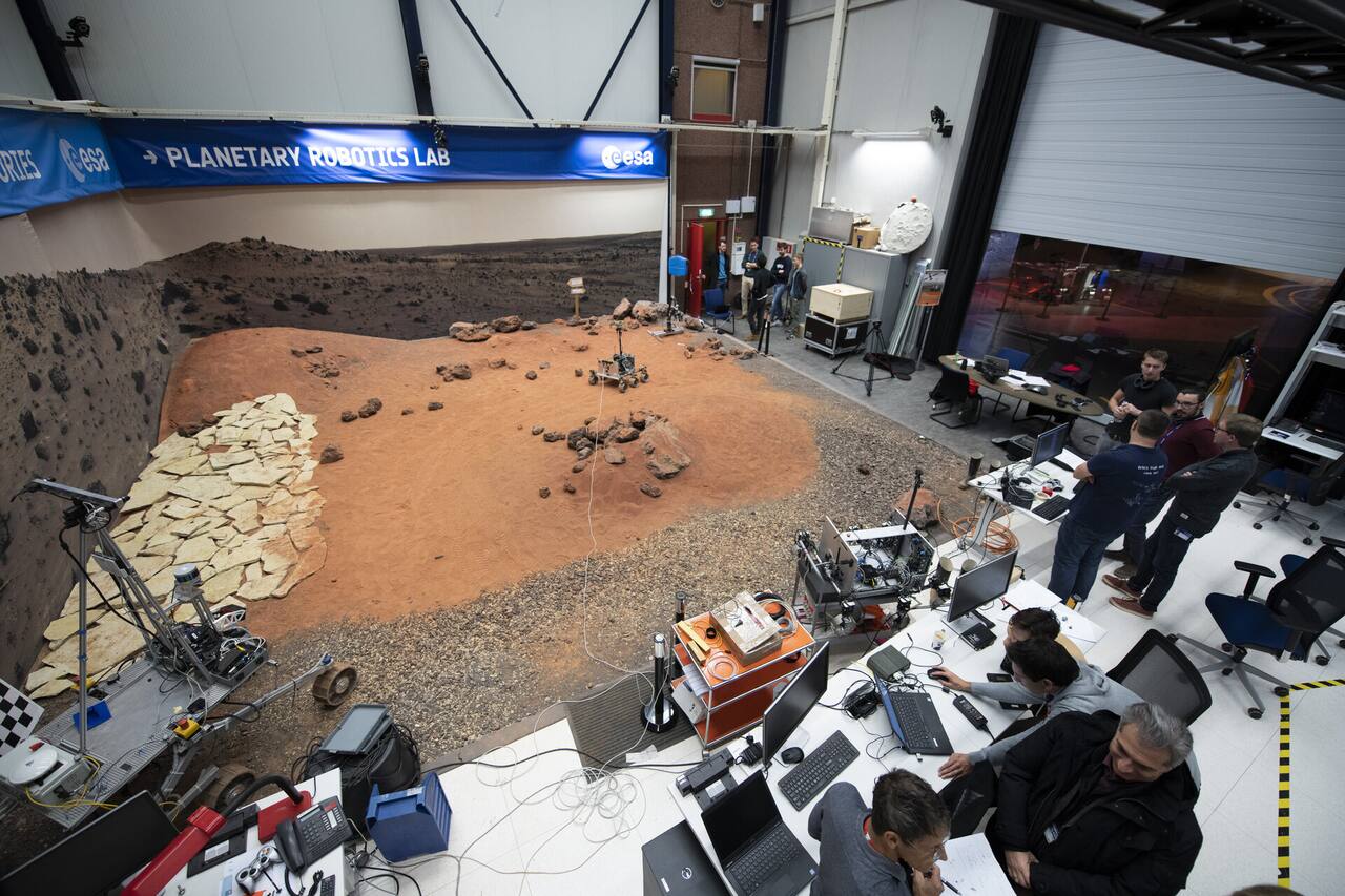 ExoTeR in the 9 by 9 m Planetary Utilization Testbed of the ESTEC Planetary Robotics Laboratory: graded sand, gravel and rock with a flagstone slope section, cameras on the overhead beam and on tripods for position ground truth, and checkerboard calibration targets at the bed edge.