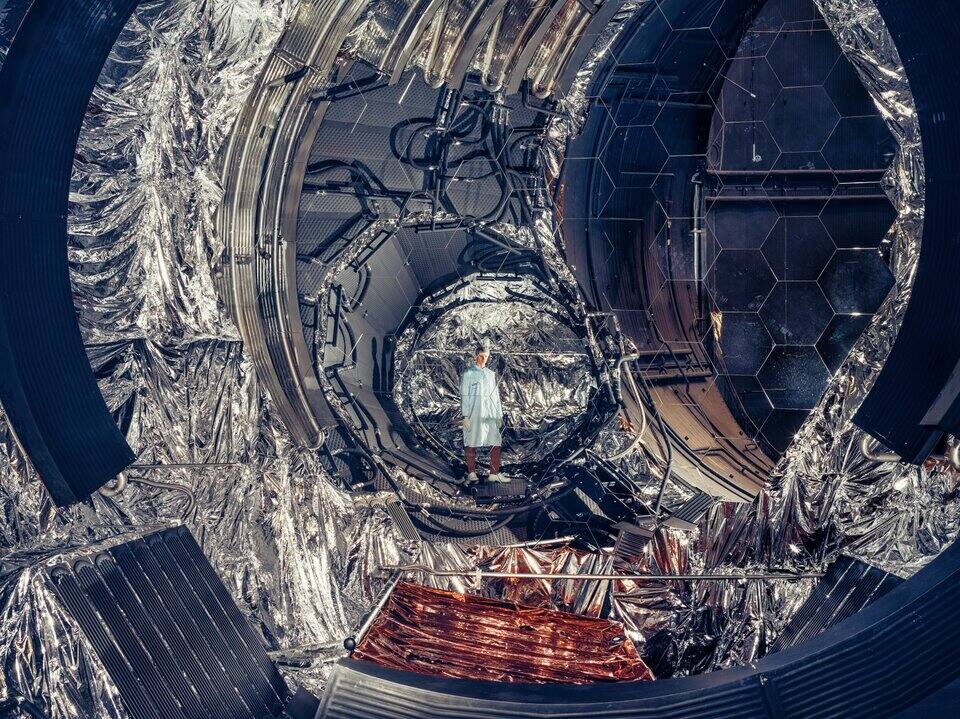 Inside the Large Space Simulator, looking down the main chamber past the hexagonal mirror array on the right and the shrouds wrapped in multi-layer insulation, with an engineer standing on the specimen platform for scale.