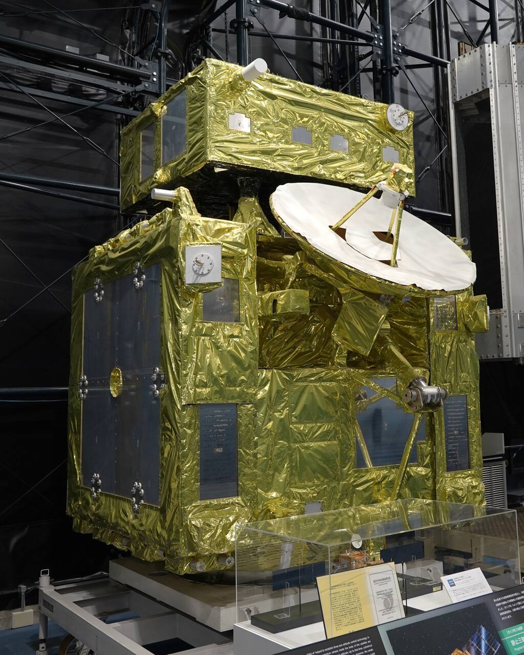 The ETS-VII display model in the Space Dome at JAXA Tsukuba Space Center, showing the launch configuration in which the two spacecraft flew mated: the smaller target Orihime on top of the larger chaser Hikoboshi, which carries the deployable high-gain antenna. This is a museum display article, not flight hardware, and the manipulator is not represented on it