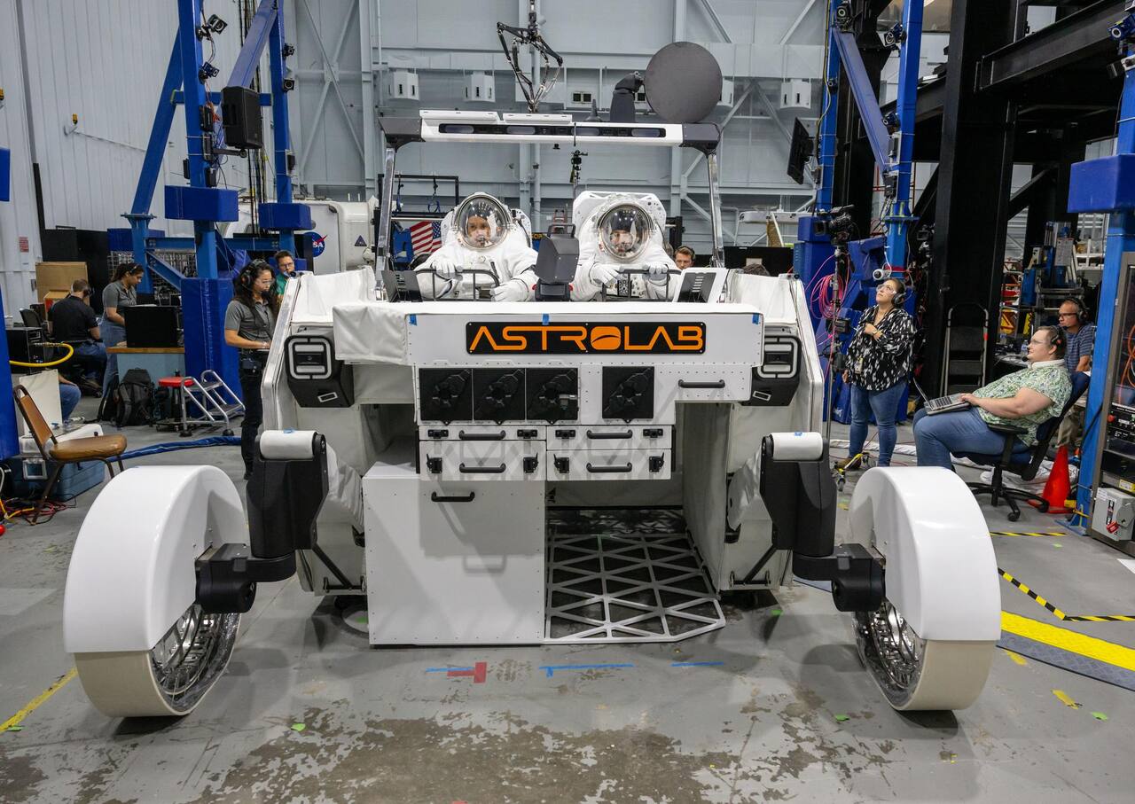 Astrolab FLEX crewed configuration during suited evaluation at NASA JSC, October 2024, seen from the front with two suited operators at the crew station. Each of the four wheels is carried on an articulating limb that sets chassis ground clearance independently; the gimballed high-gain dish is mounted above the crew station on the mast that also carries the forward camera and lighting bar, and the front face of the chassis carries tool and sample stowage drawers