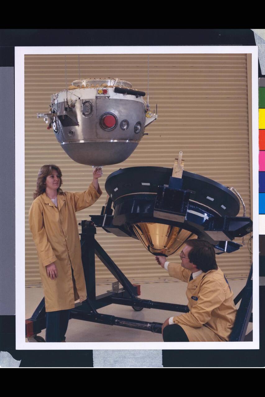 The Galileo probe descent module lifted clear of its aeroshell during assembly. The split visible here is the probe's whole architecture: the descent module above carries every instrument and every support subsystem behind the ports in its shell, and the aeroshell below, held on its handling frame, is the forward heat shield and backshell that protect it through cruise, coast and entry. The pale conical interior of the forward heat shield is exposed