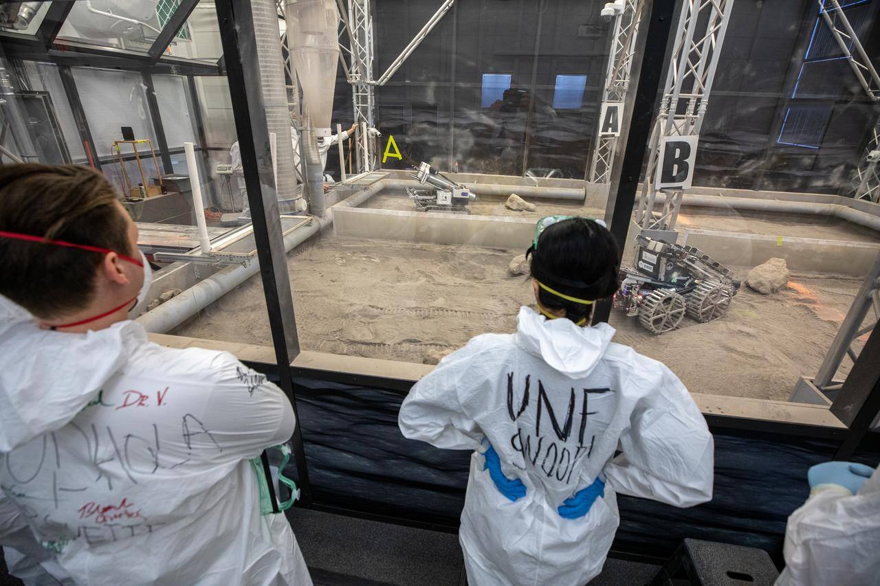 The Artemis Arena seen from the observation window: the glazed enclosure that seals the bin, the cyclone of the extraction plant that feeds the perimeter dust suppression duct, and two excavation vehicles working the simulant bed inside during a competition run.