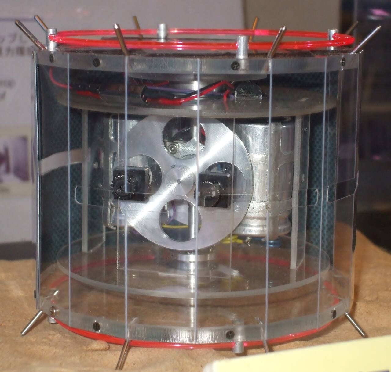 Cutaway model of MINERVA on display at ISAS Sagamihara, with a transparent shell substituted for the solar-celled hexadecagonal skin. The body is 120 mm across and 100 mm high. The internal torquer assembly at center is the entire mobility system: spinning and braking the flywheel reacts against the body to throw the rover off the surface, and the two black units flanking it are cameras. The pins projecting through the rim are the surface contact and thermal sensors
