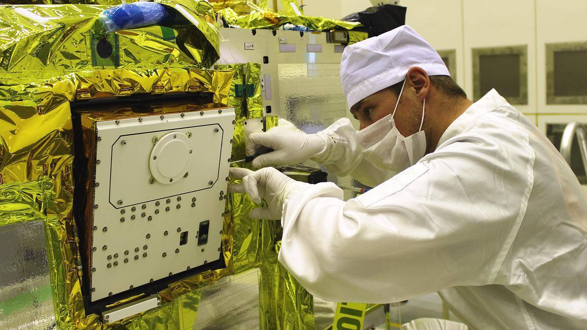MASCOT flight hardware being closed out in the cleanroom before delivery. The 30 by 30 by 20 cm box is a carbon fiber laminate structure only 0.125 mm thick, wrapped in multi-layer insulation; the white panel is the instrument face, carrying the circular aperture of the MARA thermal infrared radiometer and the camera and magnetometer ports. All instruments look one way, which is why the internal tungsten swing arm has to act as both the uprighting mechanism and the hopping actuator