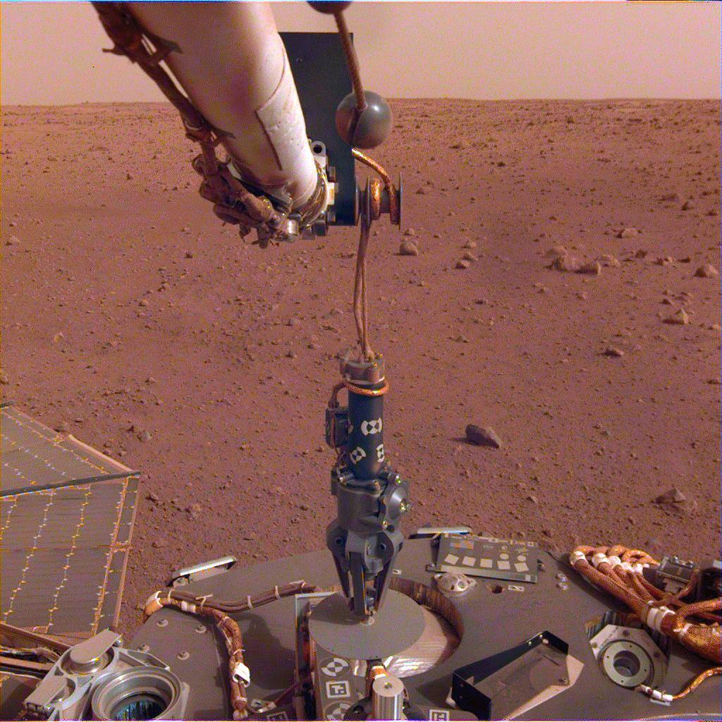 The InSight robotic arm lowering the HP3 support structure off the lander deck on 12 February 2019, the grapple still engaged on the instrument's hook. The mole descends through the tube at the center of the structure, which also carries the tether and its length reel; the mole itself is inside the tube and not visible