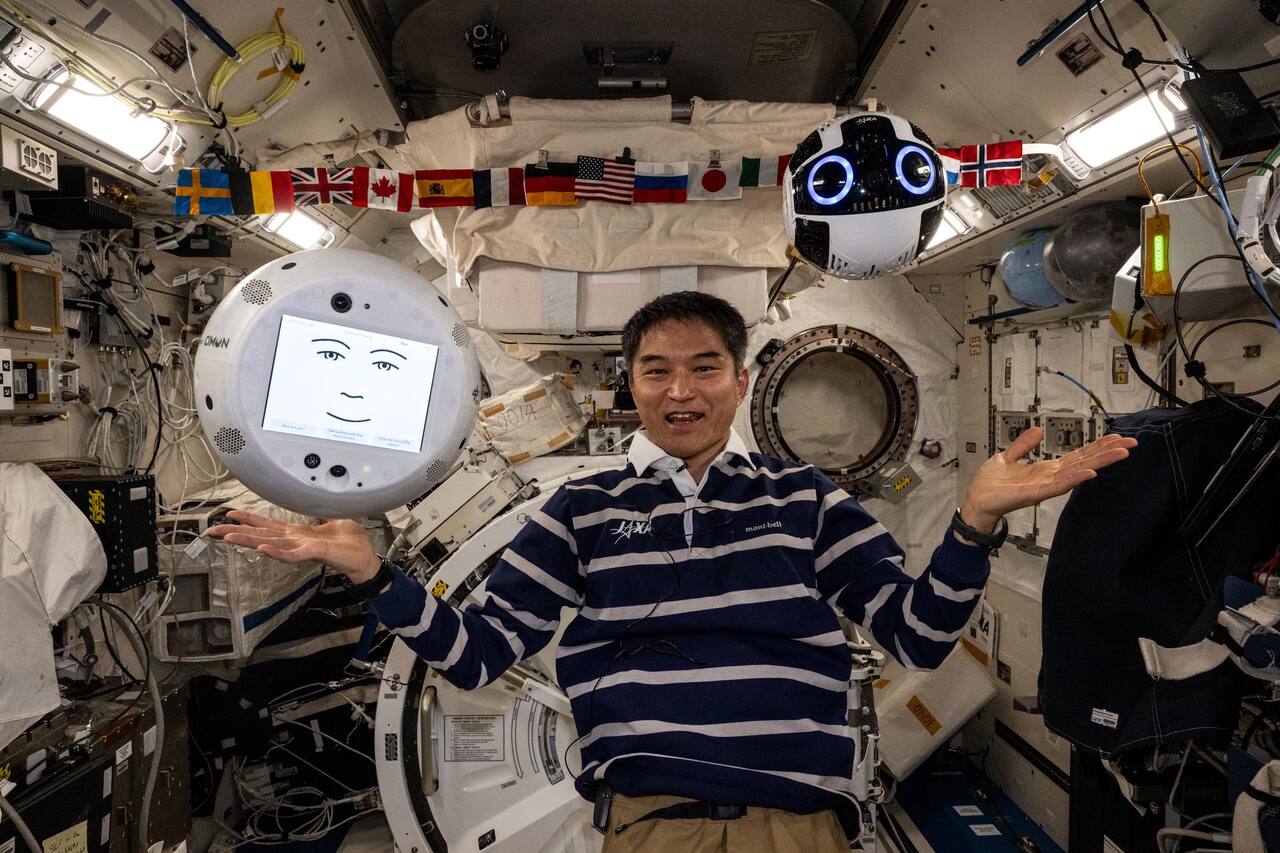 Int-Ball2, upper right, free-flying in the Japanese Experiment Module on 29 July 2025 alongside CIMON-2 at left, with JAXA astronaut Takuya Onishi between them. The 20 cm sphere carries the stereo pair used for visual-inertial SLAM behind the two illuminated ring lights, with the fan intakes and exhausts appearing as the dark perforated bands around the body. Both robots run ROS, which is what allowed them to exchange data directly in the 2025 ICHIBAN activity