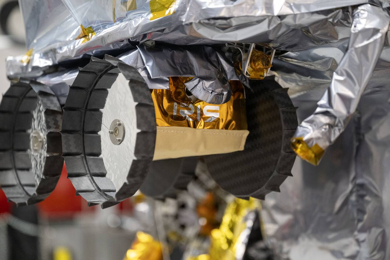 Iris secured to the Peregrine lander in the Astrobotic clean room in Pittsburgh, October 2023. The bottlecap-shaped wheels are multi-ply carbon fiber with molded grousers around the rim and a flat spoked disc face; the chassis behind them is a carbon fiber monocoque wrapped in multi-layer insulation, with gold Kapton over the body