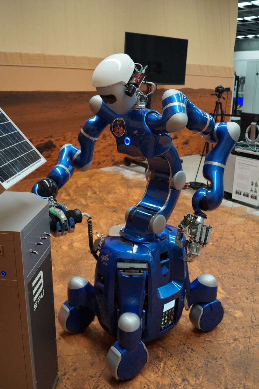 Rollin' Justin servicing a Smart Payload Unit in the Solex analogue terrain during the METERON SUPVIS Justin campaign, wearing the experiment patch on its chest. The two seven-degree-of-freedom lightweight arms terminate in twelve-degree-of-freedom hands, the right one open to show the finger drives; the head carries the stereo cameras, and the four wheel units of the omnidirectional platform are in the extended footprint. This is the ground robot that ISS crew commanded through a tablet