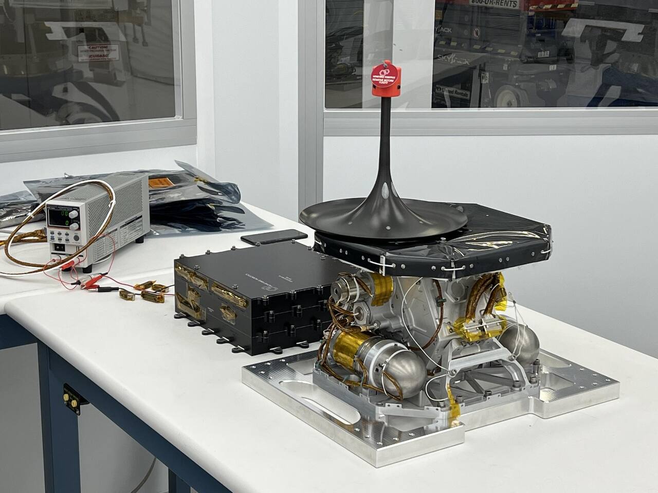 The LISTER flight mechanism on the bench before integration, with a remove-before-flight tag on the boom exit cone at the top. The black cone is the aperture the coiled stainless tubing is driven through as it is spooled out and yielded straight; the two nitrogen tanks sit inside the frame beneath it, the instrument electronics box is at the left, and the whole assembly bolts to the lander through the machined plate underneath