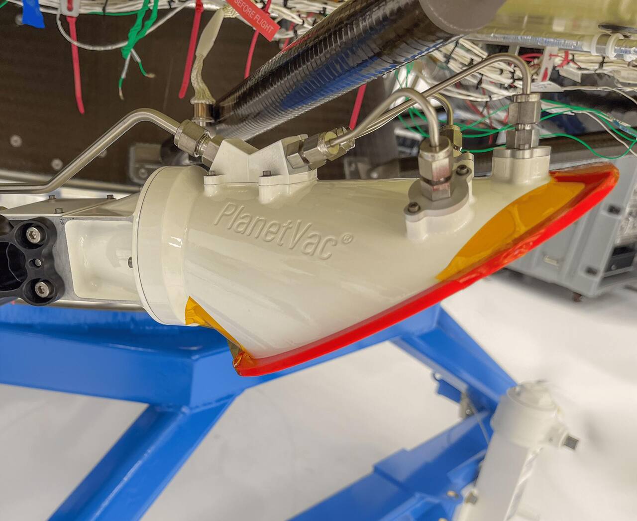 The Lunar PlanetVac sampling head mounted at the end of Firefly's Sample Access Arm beneath the Blue Ghost lander before flight, with the open mouth of the hood taped over for protection. Two rigid gas supply lines enter the top of the hood and the flexible sample transfer hose leaves it to the upper left, running up the arm to the collection system on the lander mid-deck