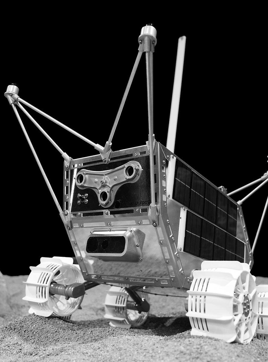 MAPP flight-configuration rover on regolith simulant. The chassis is a machined aluminum box with a body-mounted solar panel on the starboard face, a stereo navigation camera pair in a housing on the forward face, a whip antenna and two mast-mounted cameras on struts above the deck, and four independently driven wheels with open spoked hubs and cleated treads