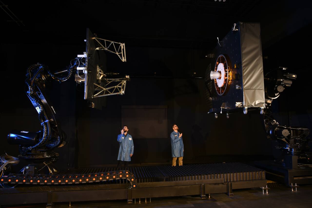 SpaceLogistics proximity operations laboratory. Two industrial robots fly full-scale articles against each other on a rail to reproduce the terminal approach. The article at right presents the client interface MEV actually uses: the zenith-facing liquid apogee engine nozzle, the white cone, inside the launch adapter ring, the pair of features present on about 80 percent of GEO satellites. The article at left carries the MEV docking mechanism with its stanchion truss extended toward that nozzle