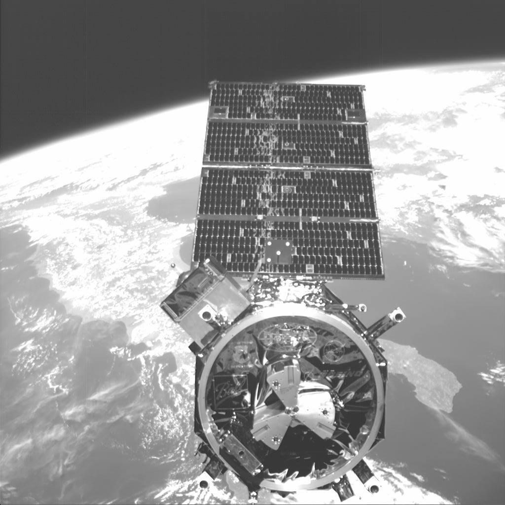 NEXTSat photographed from ASTRO during free flight in 2007, with the passive half of the docking interface facing the camera. The ring, specified at under 46 cm outer diameter, carries the three-point kinematic mount that ASTRO's motor-driven linkages close on; the electrical and fluid couplings sit inside it, so mating the structure mates the utilities. Nine mate and demate cycles were flown through this interface