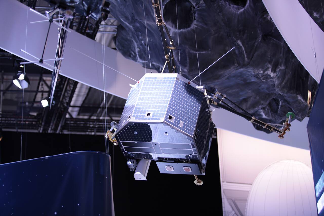 A full-scale Philae model suspended under a comet nucleus mock-up. The hexagonal body, about 0.7 by 0.7 by 0.9 m, is covered on all six sides by the solar panels that carried 1224 cells; the tripod landing gear with its three foot pads spans about 2.3 m across the feet, and the balcony structure at the base holds instrument apertures. None of the four hold-down systems that were to have anchored these feet worked at touchdown