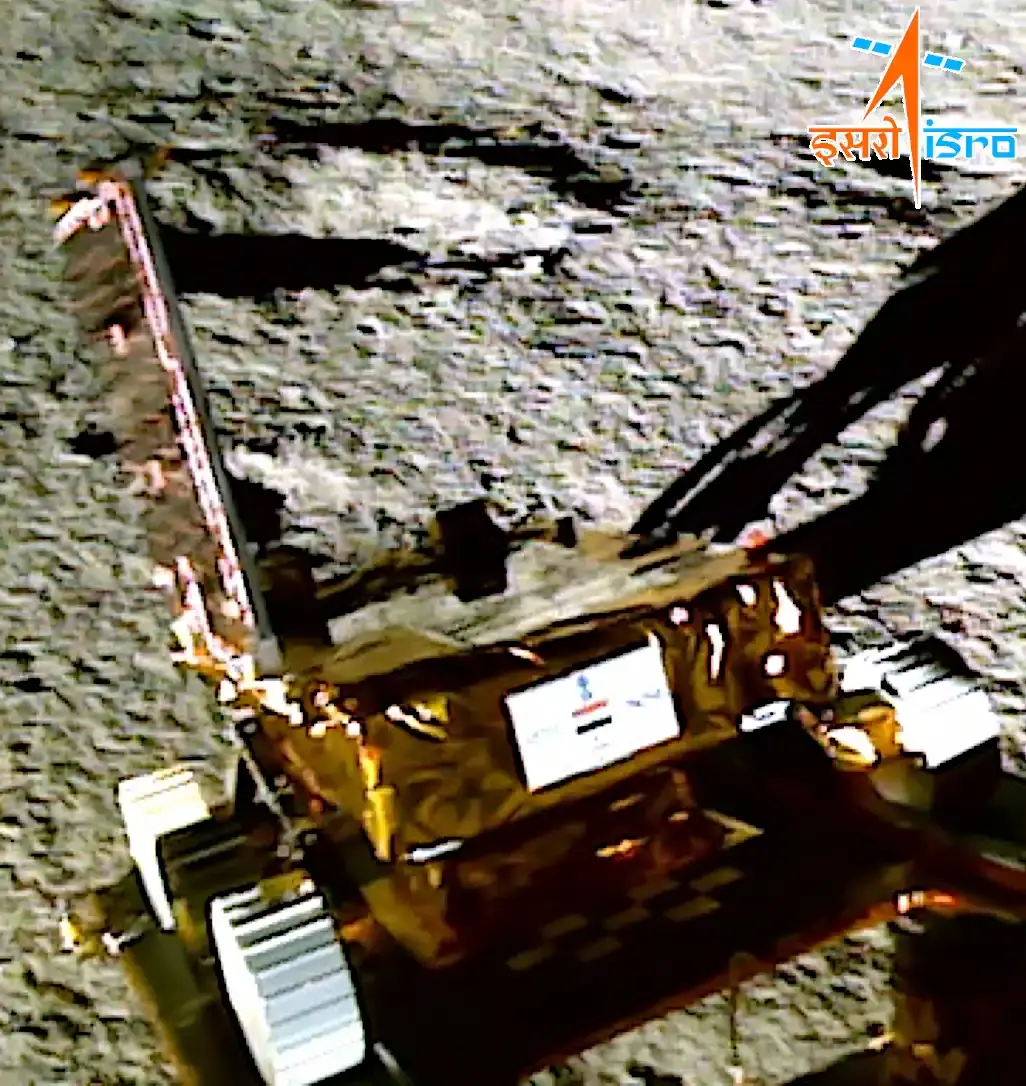 Pragyan on the lunar surface after ramp-down from Vikram, August 2023. The gold multilayer-insulated chassis carries the solar panel on a raised hinge at left, one grousered wheel is visible at lower left, and the body panel carries the Indian national emblem