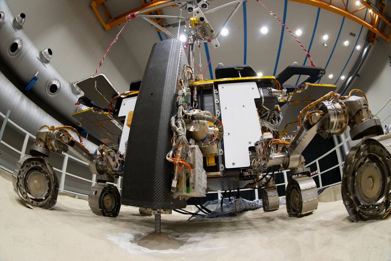 Rosalind Franklin ground test model during the March 2023 drilling campaign in Italy, in which the drill reached 1.7 m into Mars-analog ground and returned samples. The drill box stands vertically ahead of the front face with the string in the soil, and the triple-bogie suspension carries six flexible grousered wheels, each on its own steering drive and its own articulated deployment drive