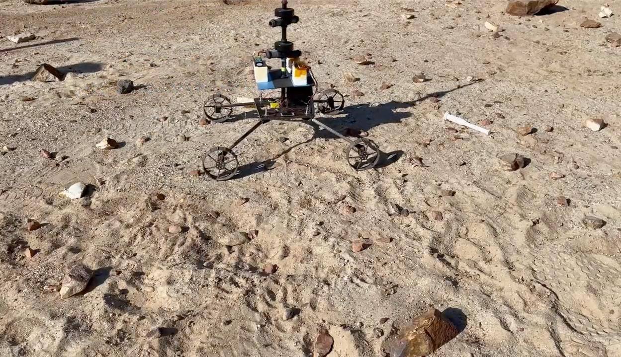 Sample Recovery Helicopter ground mobility test model driving on Mars-analog terrain, December 2022. Rotor blades are not fitted. The four driven wheels carried on the landing legs are the addition that separates this design from Ingenuity, and the tracks behind the vehicle record a driven traverse rather than a landing