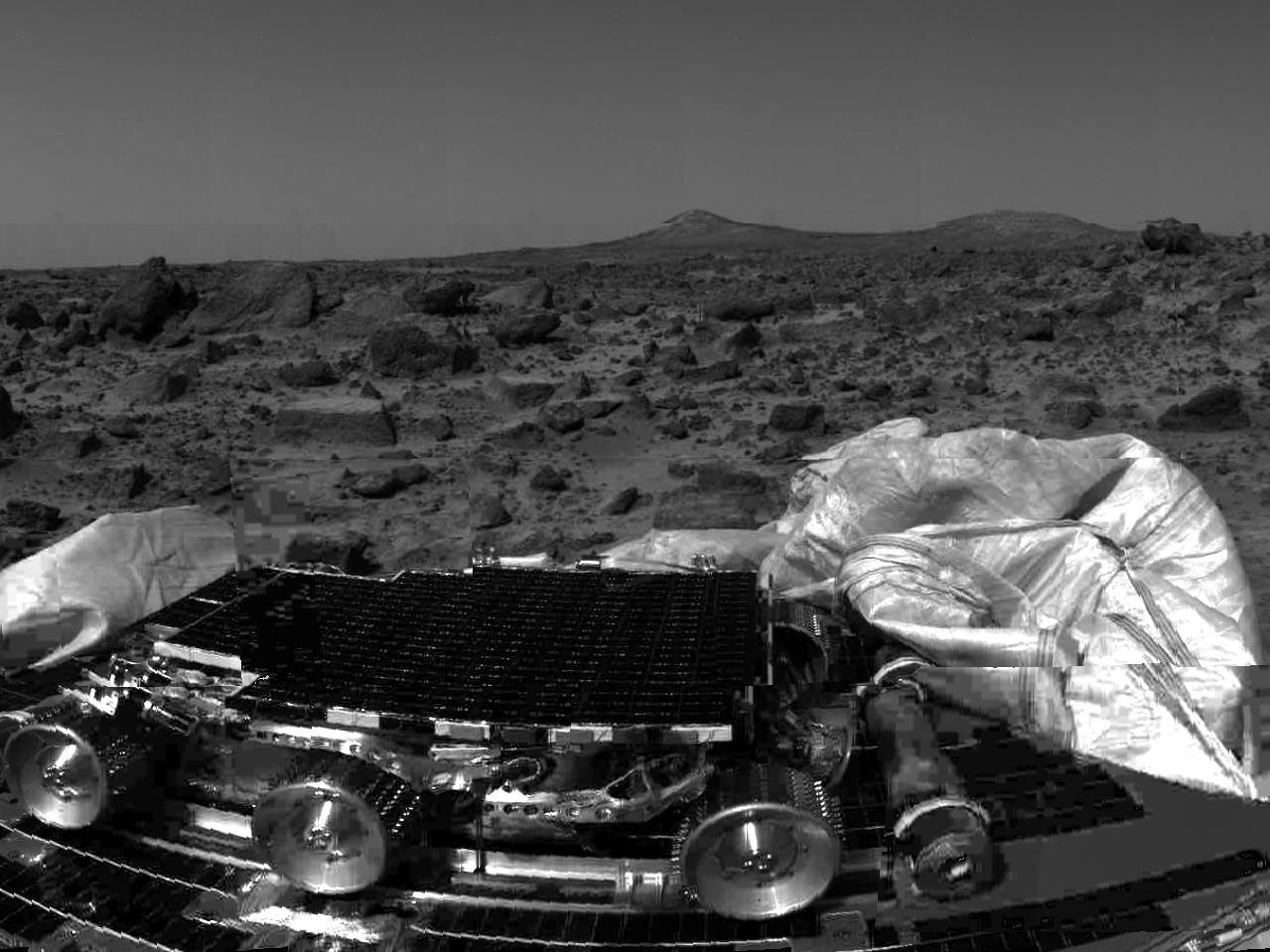 Sojourner still latched to a Pathfinder lander petal on sol 1, imaged by the Imager for Mars Pathfinder. The six wheels, the flat solar panel that covers the whole deck and the deployment arrangement on the petal are visible, with a deflated airbag in the foreground