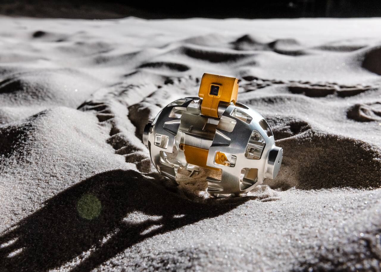 SORA-Q in its deployed running configuration on a regolith simulant bed. The two hemispherical shells have opened out into wheels, the yellow front camera housing stands proud of the body between them, and the tail stabilizer trails behind in the dust