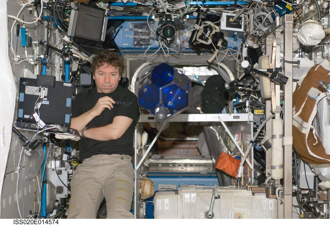 A SPHERES satellite free-flying in the ISS on 26 June 2009. The 18-sided polyhedral shell is about 20 cm across and carries the ultrasonic and infrared metrology receivers as the small circular ports distributed over its faces; the gray cylinder protruding from the lower face is the replaceable carbon dioxide tank that feeds the twelve cold gas thrusters. The satellite is unpowered by the station and runs on crew-replaced batteries