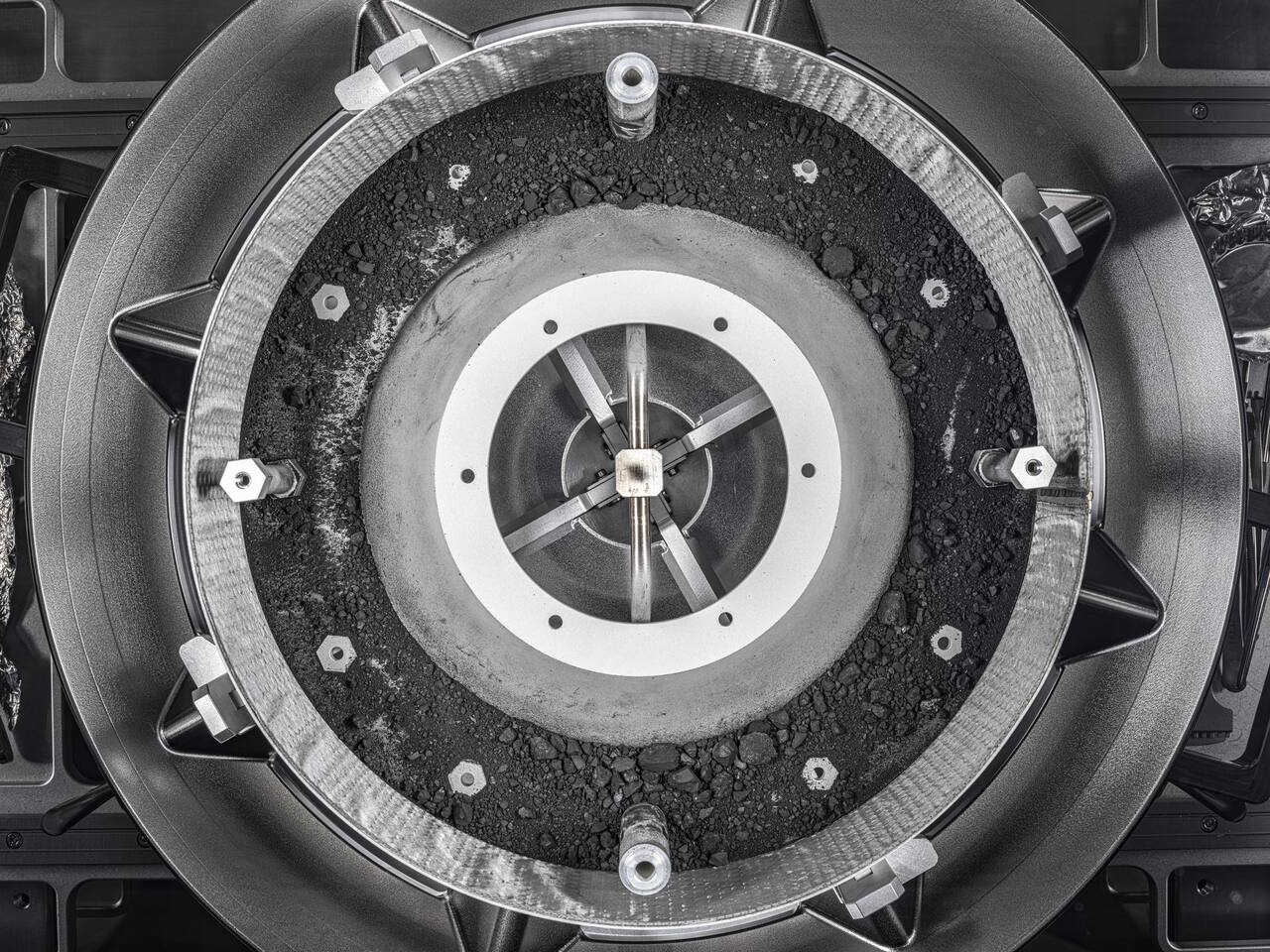 The TAGSAM head opened at NASA JSC on 17 January 2024, with Bennu material lying in the annular collection chamber. The head is about 7 cm tall on a 32 cm base plate with a 21 cm opening; the outer flange carries the stainless steel Velcro contact pad ring that captures grains from the surface, and the central hub is the nitrogen manifold that fluidized the regolith one second after contact