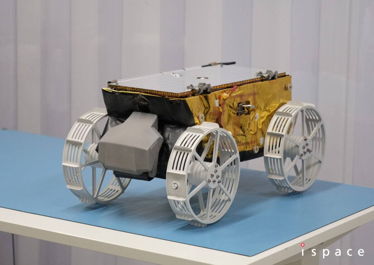 TENACIOUS flight model on completion at ispace-EUROPE in Luxembourg, July 2024. The carbon fiber reinforced plastic chassis is wrapped in gold multi-layer insulation and closed on top by a body-mounted solar panel; a whip antenna rises from the rear deck, the four wheels are rigid spoked rims with slotted circumferential grousers and no suspension, and an enclosure is mounted to the side of the chassis between the wheels