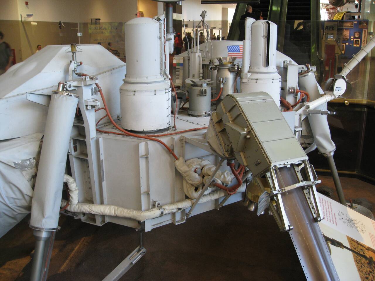 Viking lander proof test article at the National Air and Space Museum. The surface sampler acquisition assembly stands at right of center on the lander deck: the curved housing encloses the storage drum, and the flattened furlable boom element runs out of it along its guide rails. The boom forms a rigid column only once extended through those guides