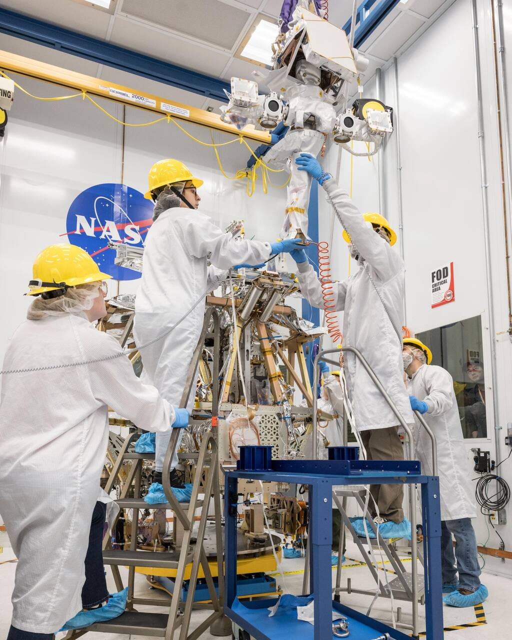The VIPER flight rover in the Johnson Space Center clean room on 12 February 2024, as the mast is lowered onto the chassis on the crane beam above. The mast head carries the stereo navigation camera pair in the two circular housings and the payload deck of the rover body is open below it