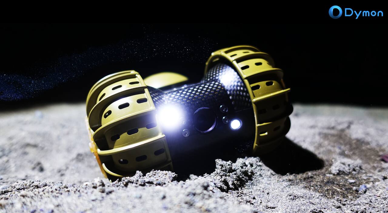 YAOKI in a manufacturer publicity image on regolith simulant. The vehicle is two large wheels on a single common axle either side of a dumbbell-shaped carbon fiber body; each wheel rim carries slotted circumferential grousers, and the camera aperture is centred on the body face between two LEDs, so the same optical assembly works whichever way up the rover comes to rest
