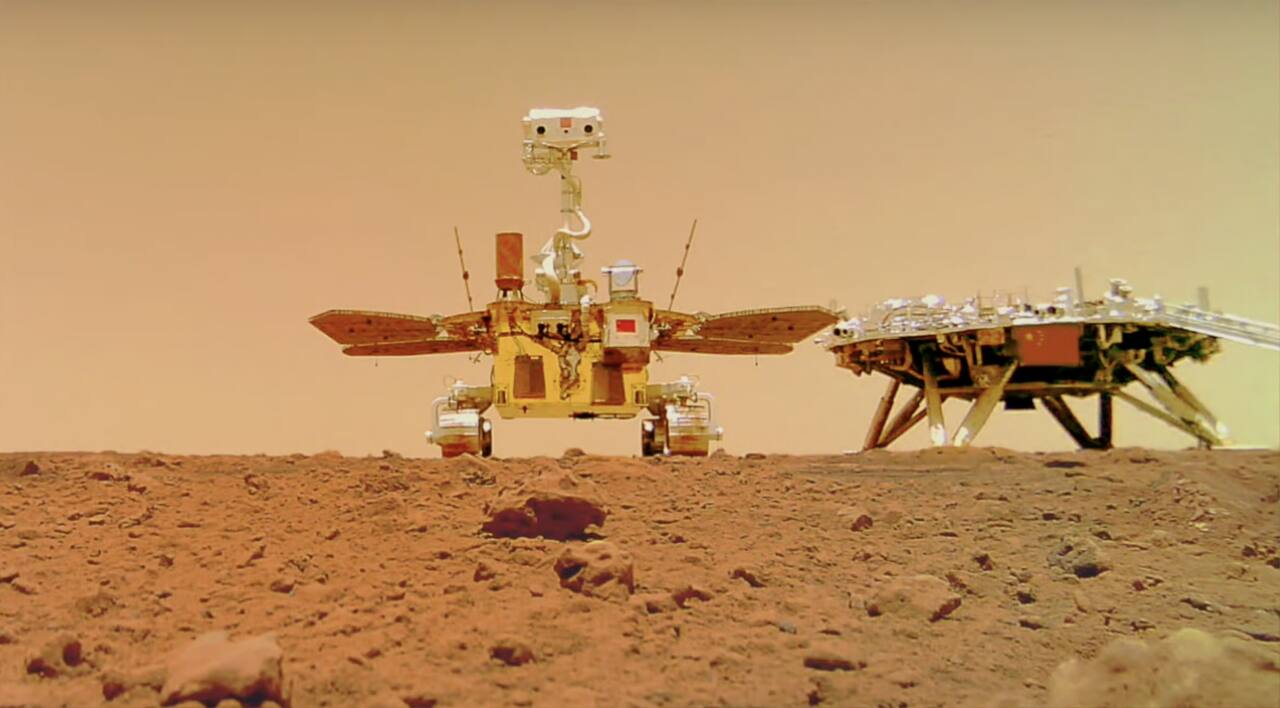 Zhurong beside the Tianwen-1 landing platform in Utopia Planitia, photographed by the deployable remote camera the rover placed on the ground and then drove away from. The six wheels, the four deployable solar panels and the mast are visible, as is the ramp the rover descended from the platform